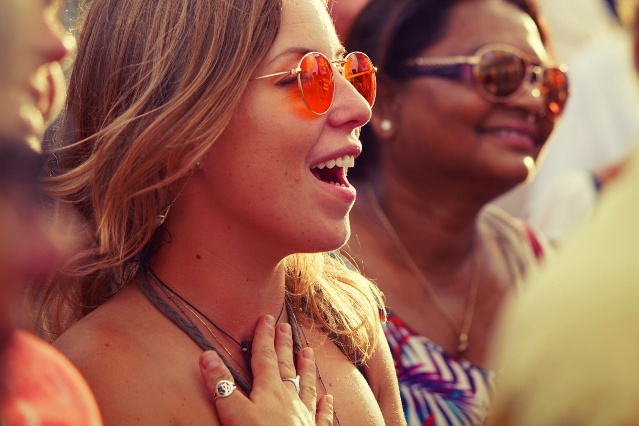 A close-up of several women outdoors in bohemian-style attire, jewelry, and orange sunglasses, being excited while looking somewhere.