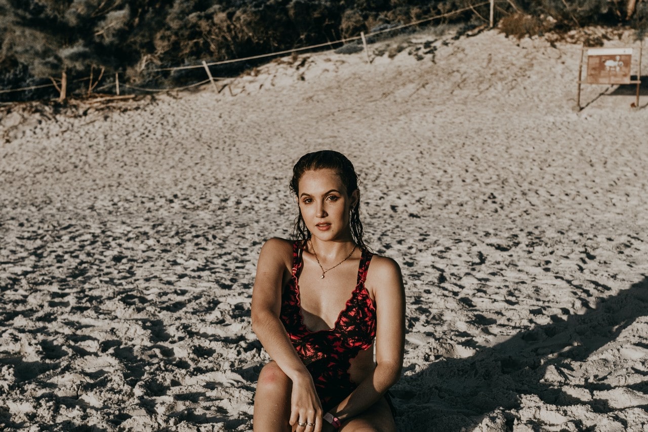 A picture of a woman in a swimsuit sitting on a sandy beach with wet hair, while looking at the camera and showcasing a yellow gold necklace.