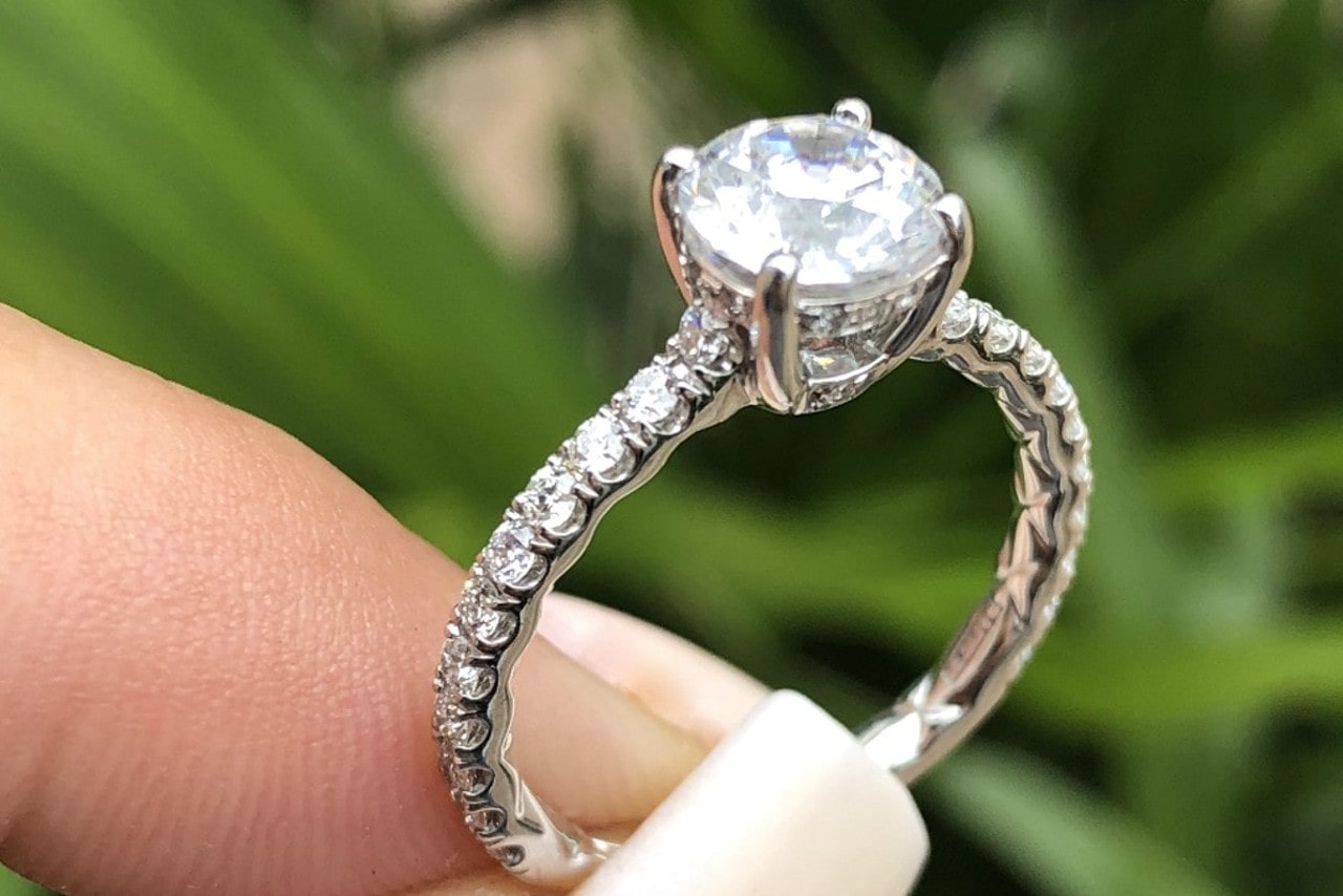 A close-up of a person holding a side-stone engagement ring with a round-cut stone.
