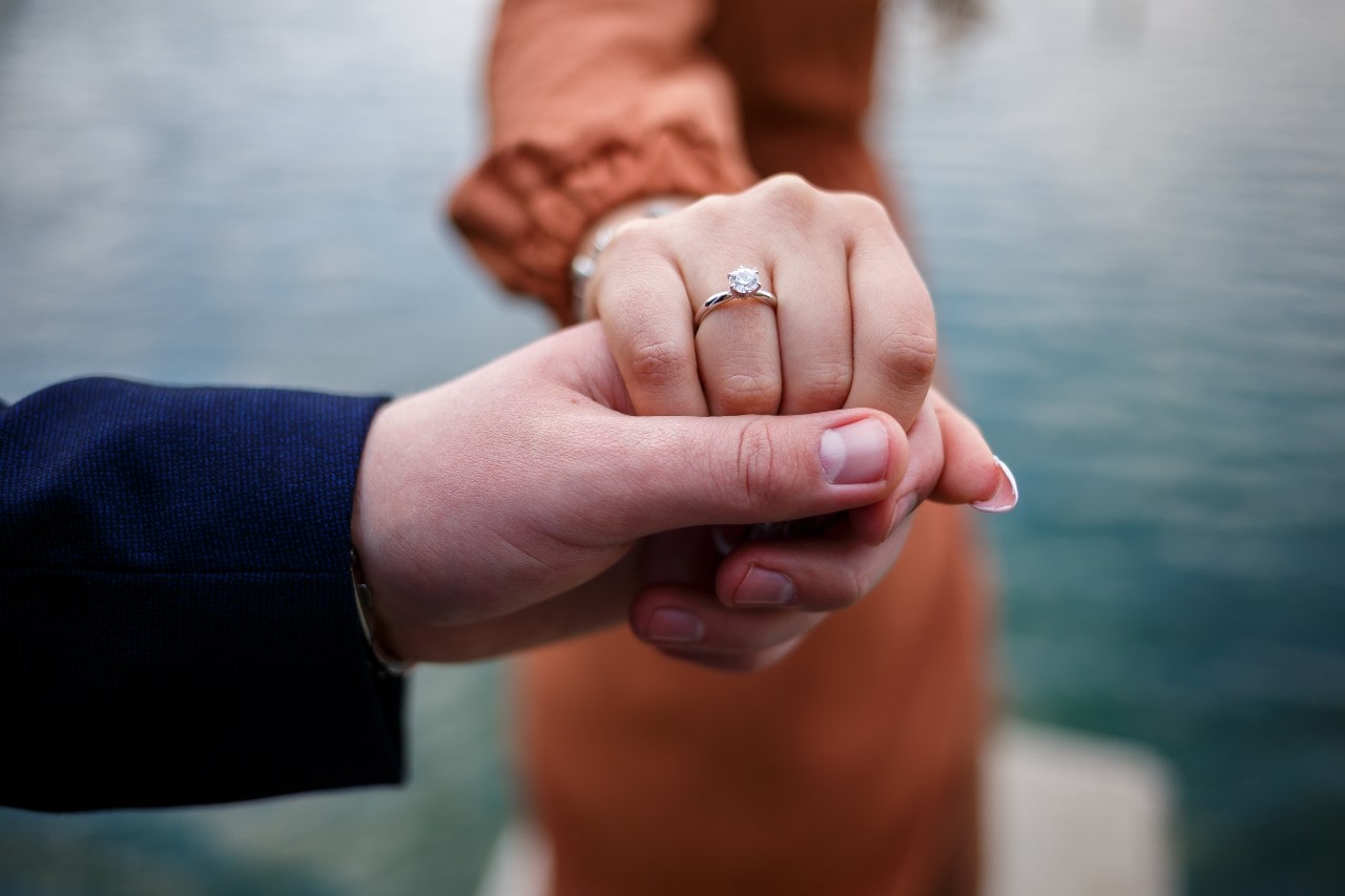 A couple holding hands on a dock, showcasing the woman&rsquo;s engagement ring.