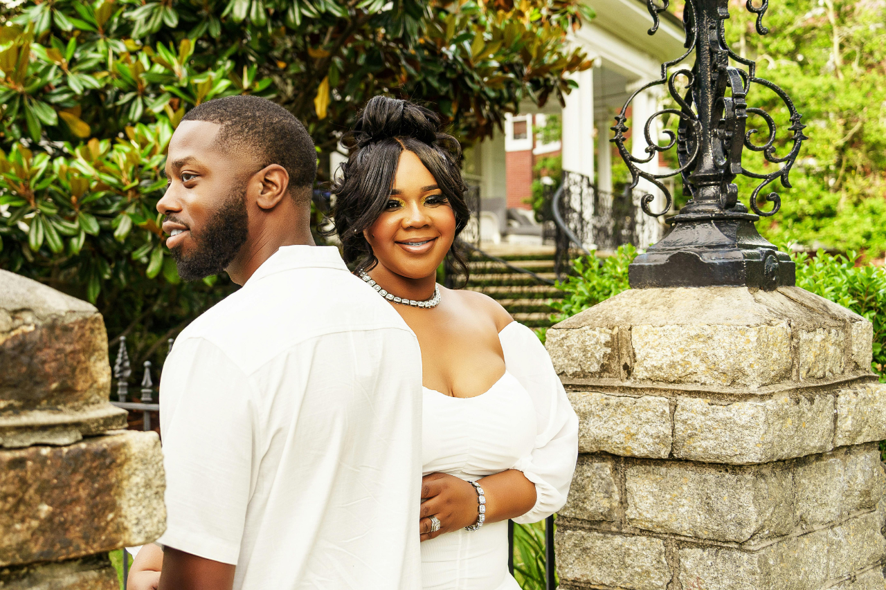 A photo of a young engaged couple in a lush New Orleans garden, both wearing white and standing between the brick columns of an iron gate.