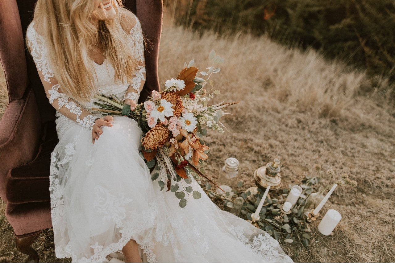 a bride sits in a chain in a fall field holding a bouquet.