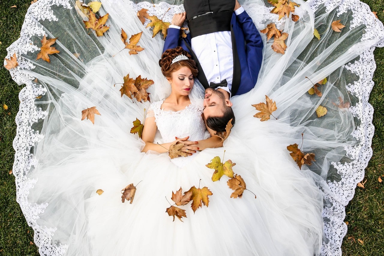 A bride and groom lay down in the grass as leaves fall on them.