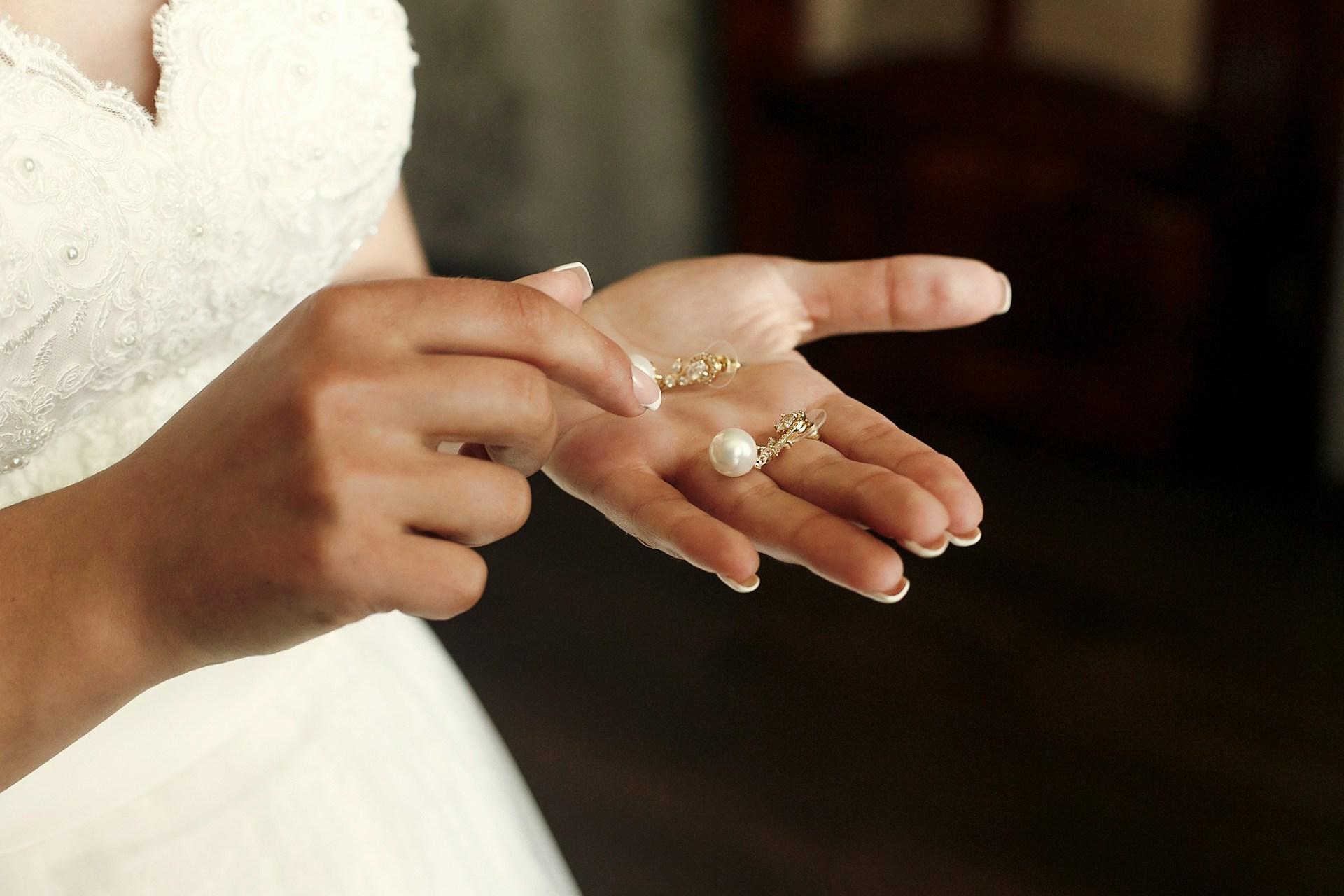 A close-up of a bride in a wedding dress holding a pair of pearl drop earrings in the palm of her hands.