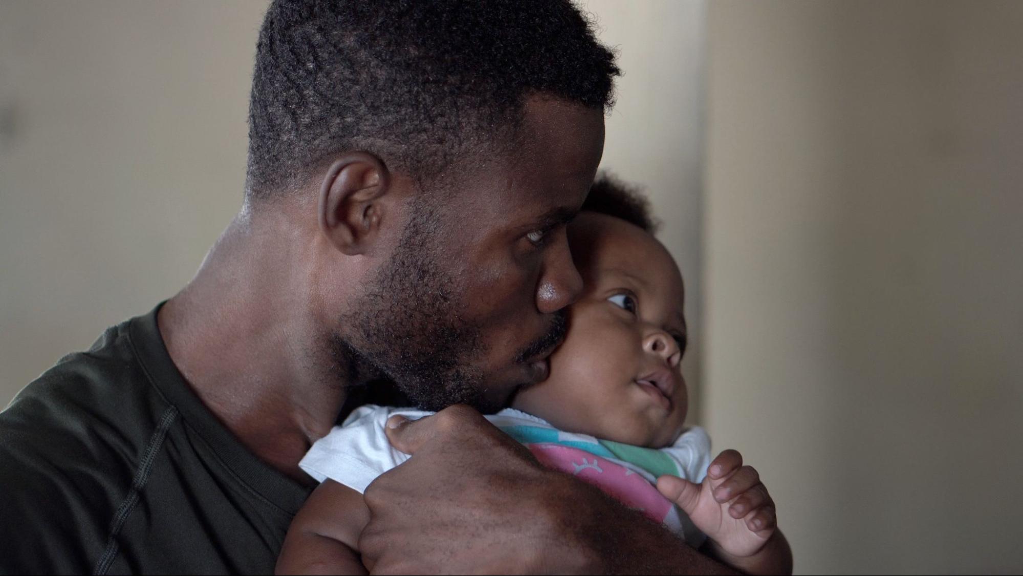 A father kissing his baby on the cheek while he holds them close to his chest.