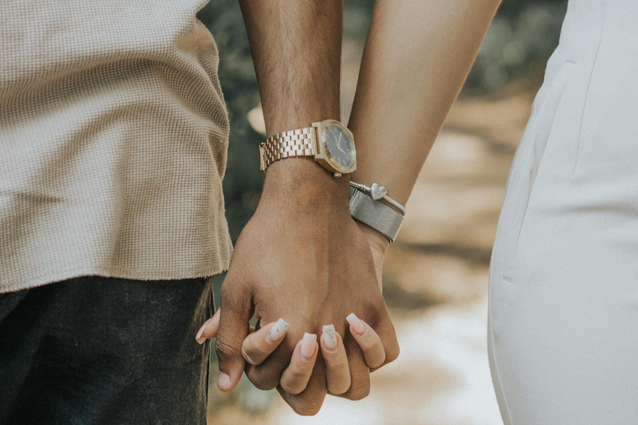 A couple's intertwining hands, showcasing a yellow gold watch and metallic bracelets.