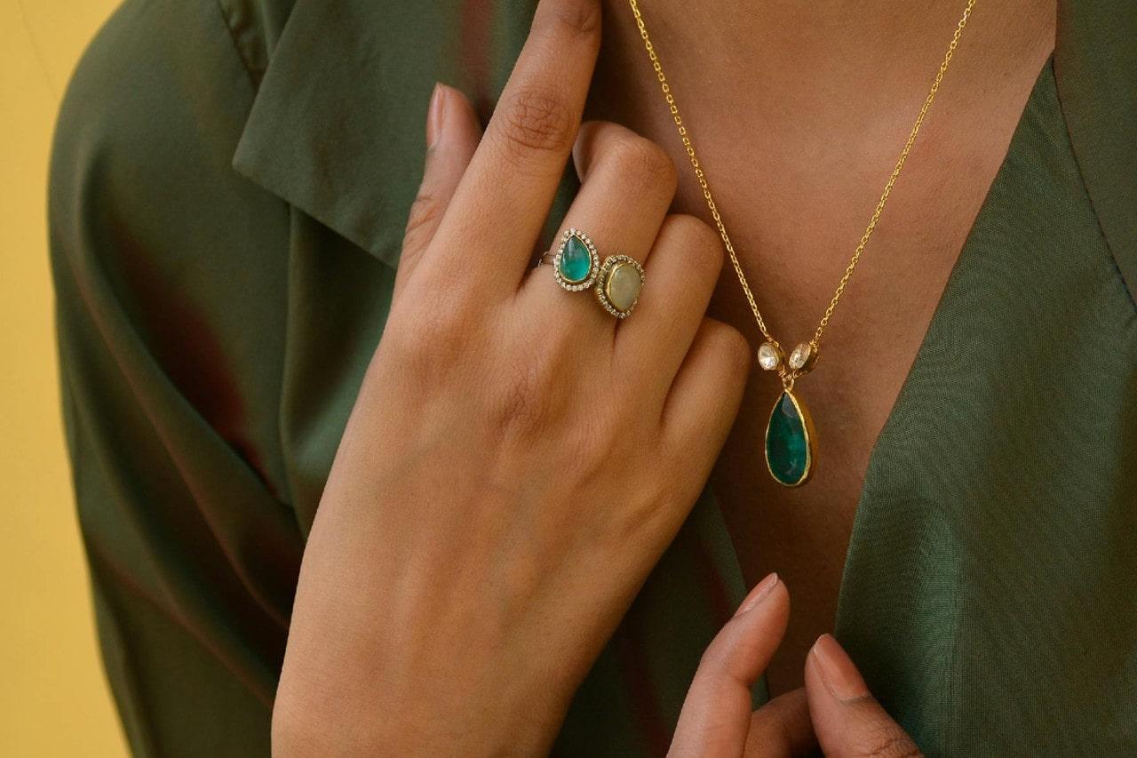 A close-up of a woman wearing a gold necklace with a teardrop-shaped green gemstone pendant and a ring featuring two pear-shaped gemstones, against a deep green blouse