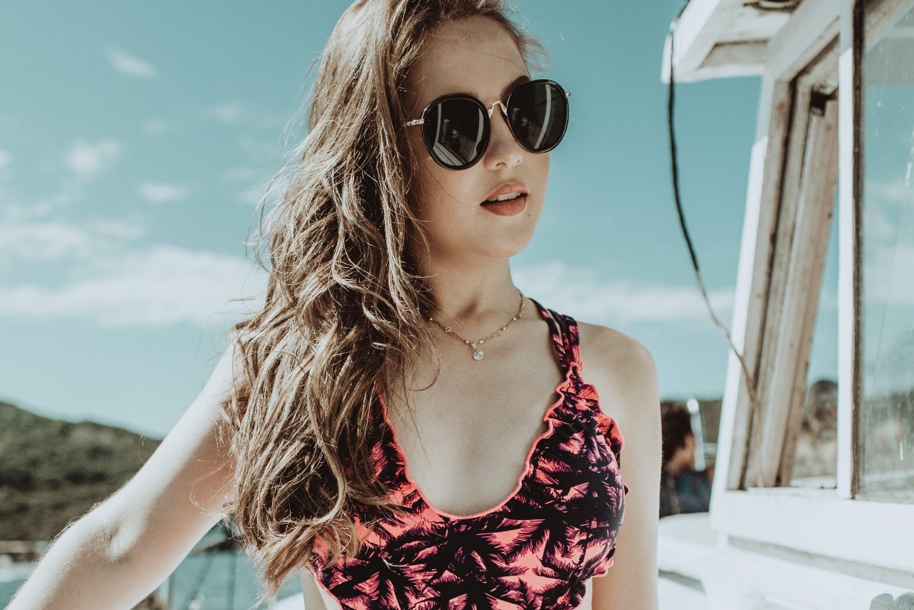 A woman in a pink palm print swimsuit with sunglasses, and yellow gold diamond necklaces stands in front of the sunlight sky.
