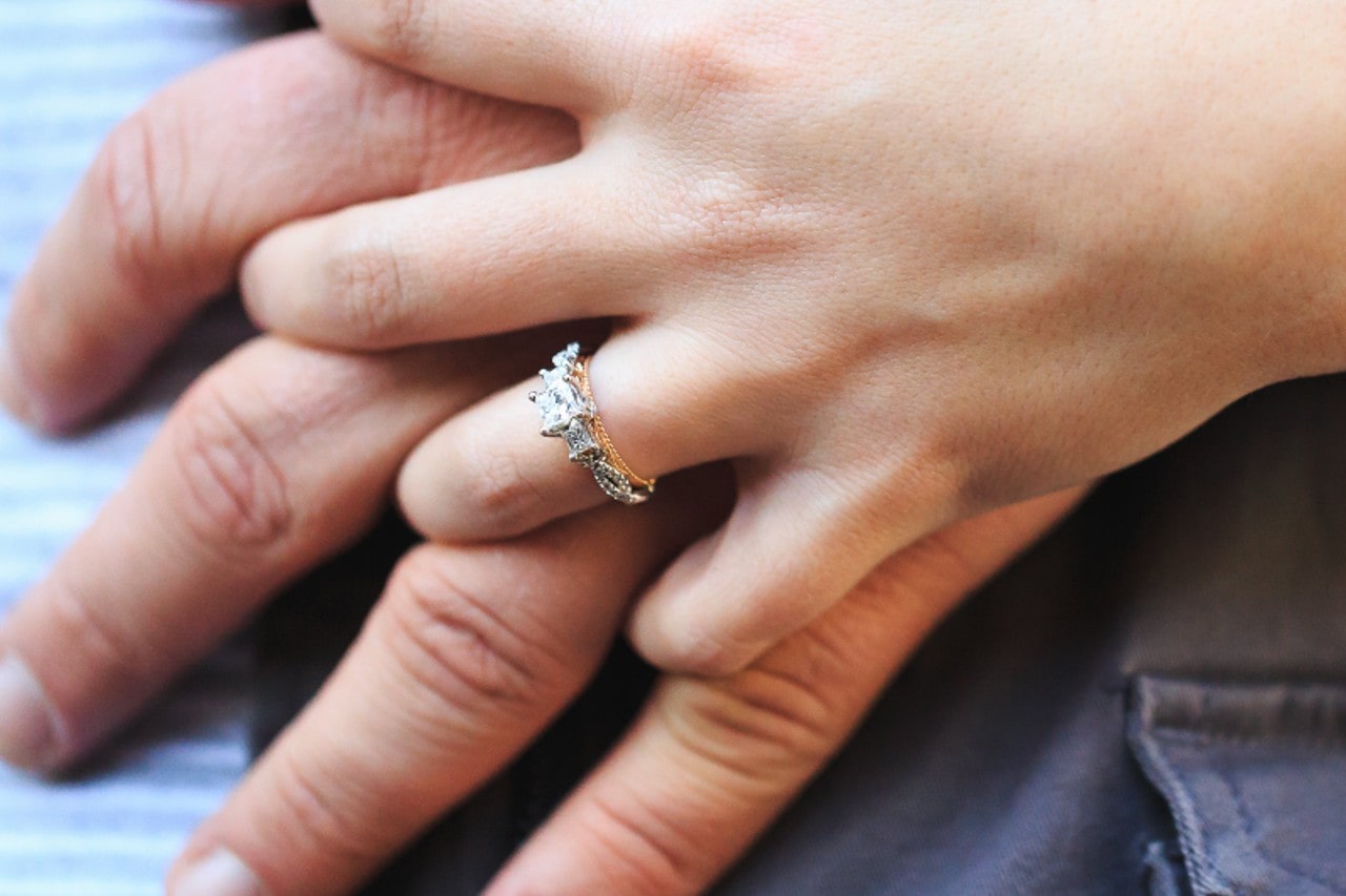 a couple’s hands intertwined, the woman’s hand donning a mixed metal, three stone engagement ring