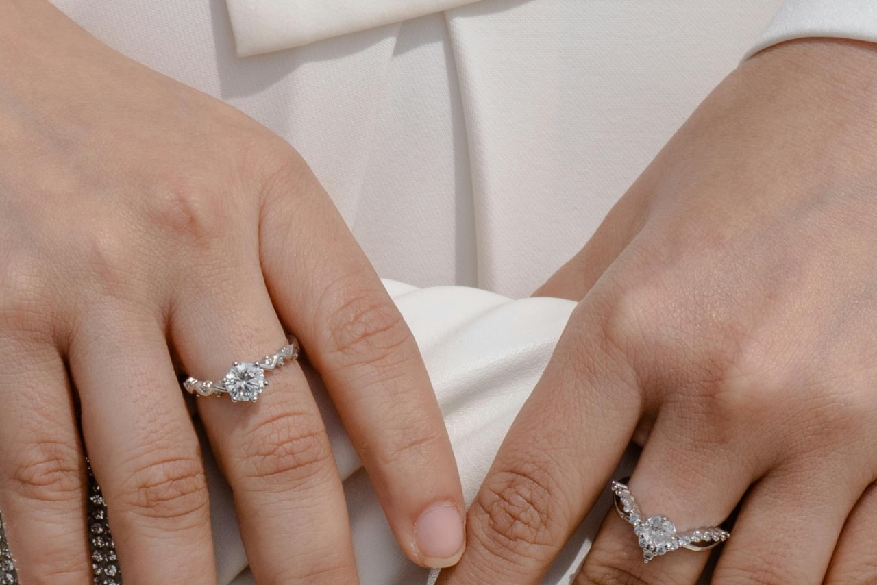 A close-up of a woman&rsquo;s hands holding a white clutch, wearing ornate diamond rings on each of her middle fingers.