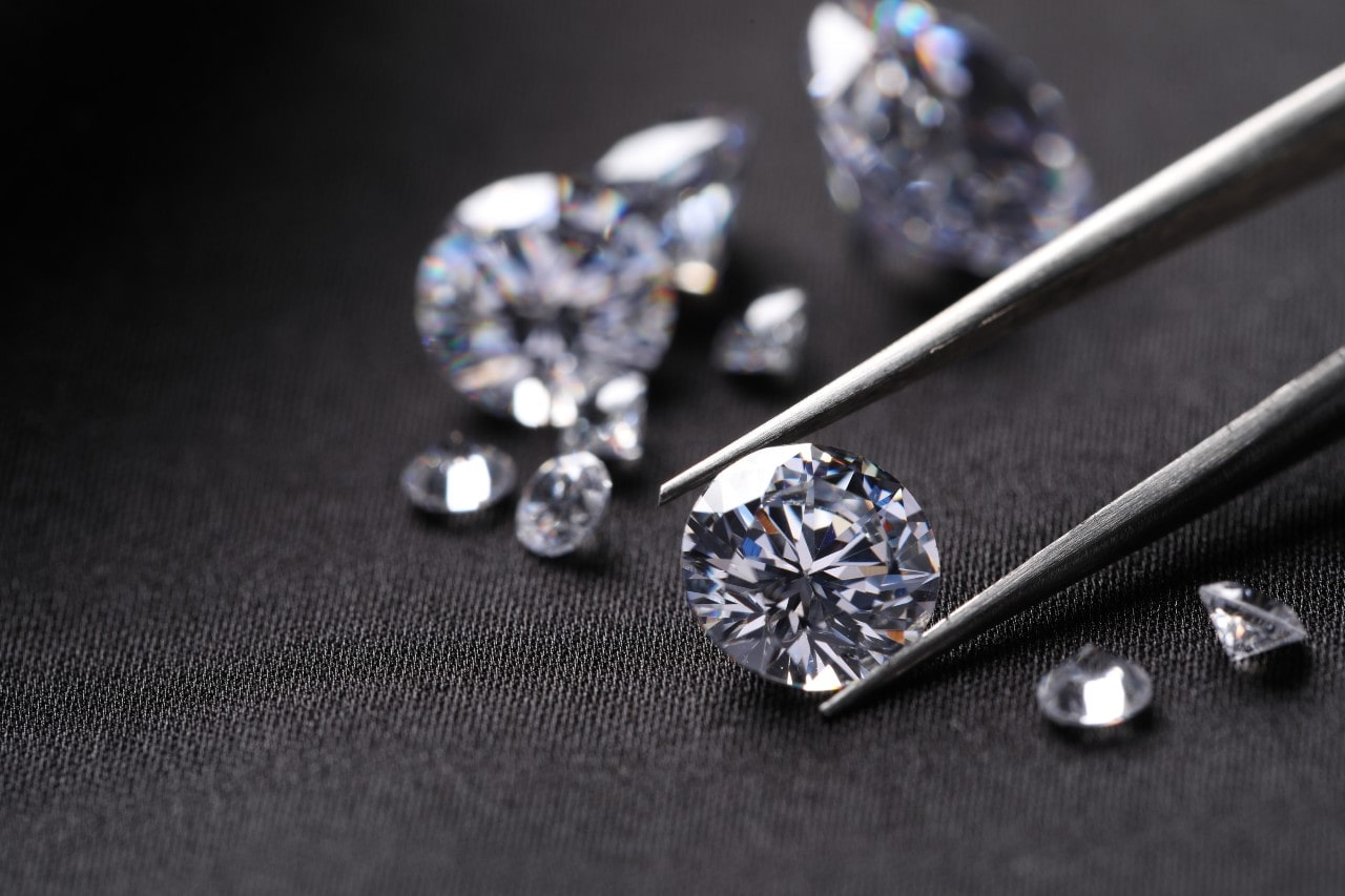 A close-up of a pair of jeweler’s tweezers gently holding a sparkling round cut diamond on a dark denim surface, with larger and smaller diamonds framing the tweezers.