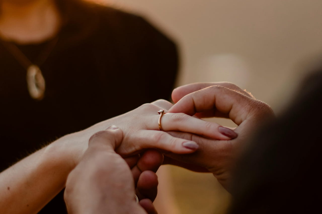 A close-up of a person gently sliding a gold engagement ring onto their partner's finger.