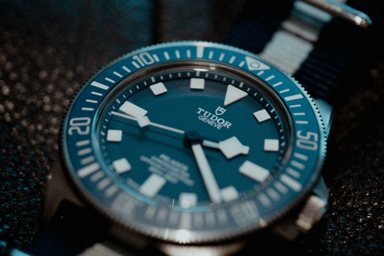 Close-up of a Tudor Genève dive watch with a blue bezel, bold indices, and a NATO strap.