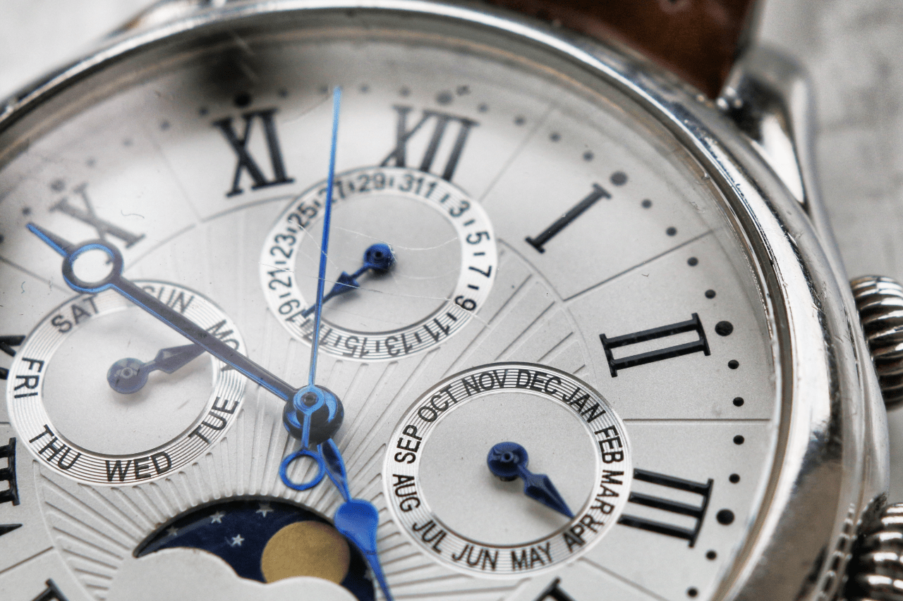 Close-up of a wristwatch with Roman numerals, blue hands, and multiple subdials showing days, months, and moon phase.