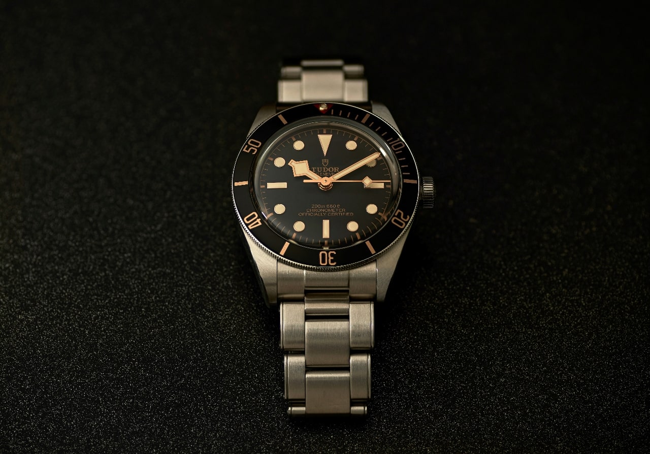 A close up of a TUDOR watch with black dial and metallic bracelet against a black textured surface.