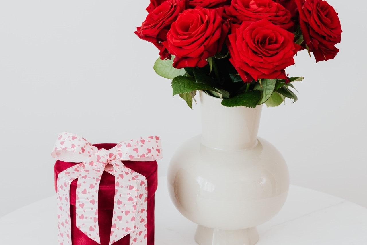 a red gift box with a pink ribbon and a white vase of red roses