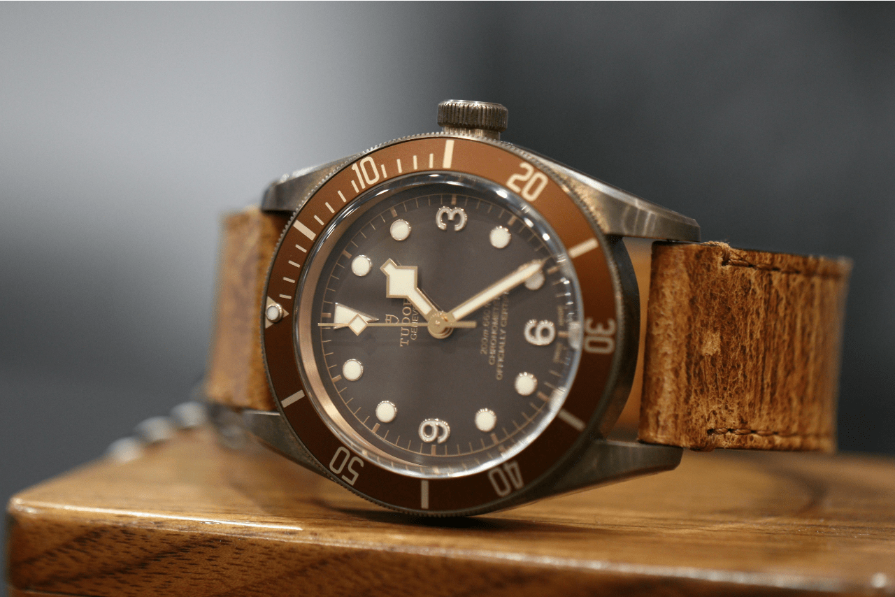 A close up of a TUDOR wristwatch with black dial and brown leather bracelet on top of the wooden surface.