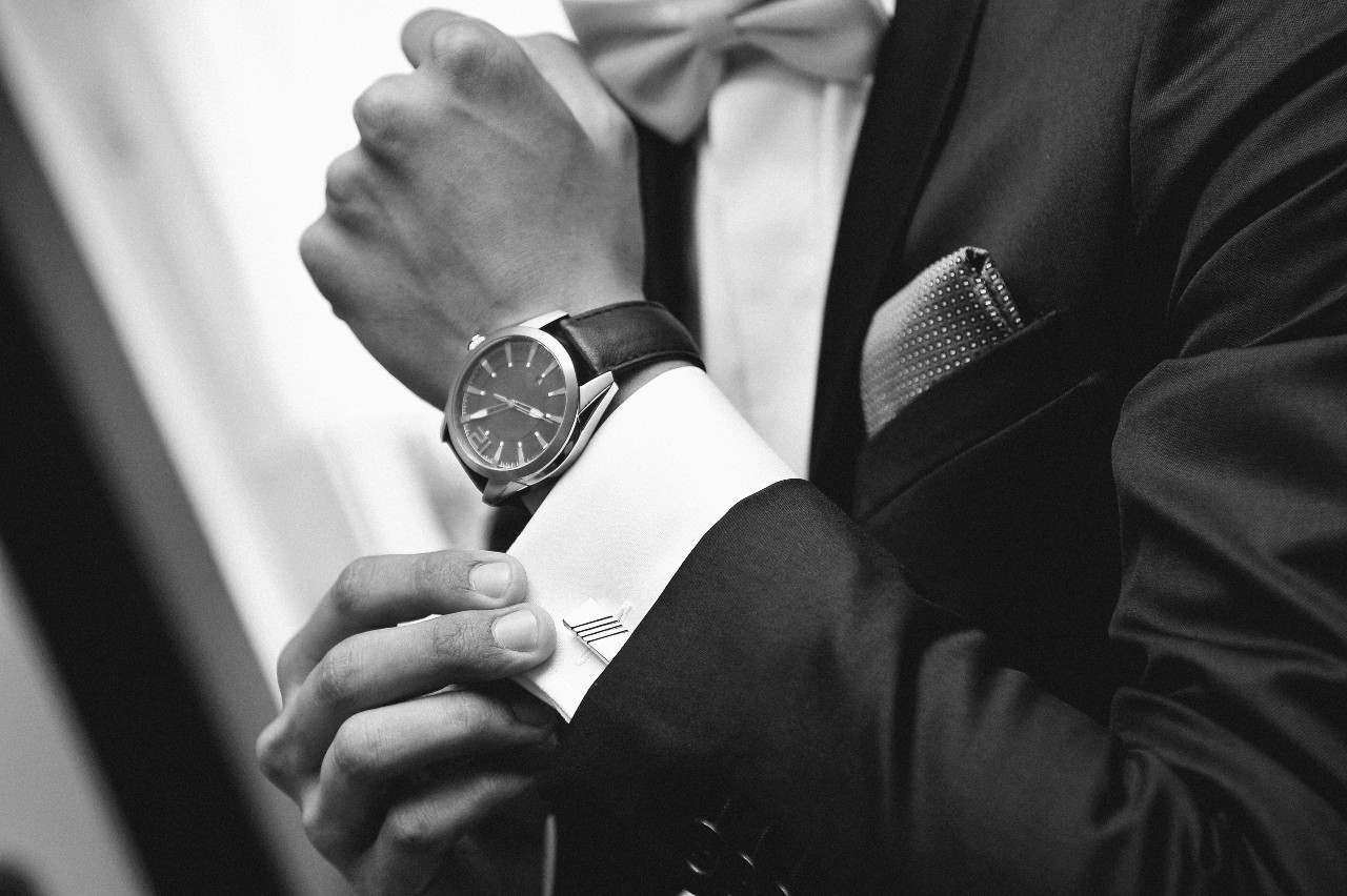 A black and white photo of a man in a suit fixing his cuffs, showcasing a wristwatch with black leather bracelet.