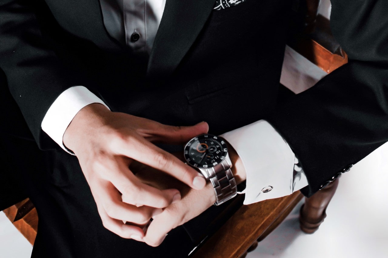 A close up of a man in a suit fixing his metallic wristwatch with black dial while sitting on a wooden chair