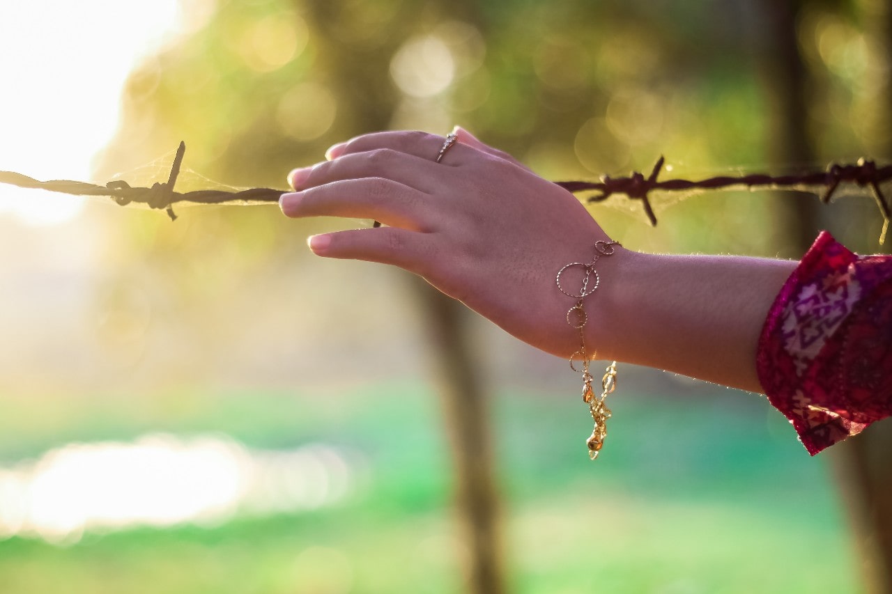 a woman touches a barbed wire fence wearing a gold chain bracelet.