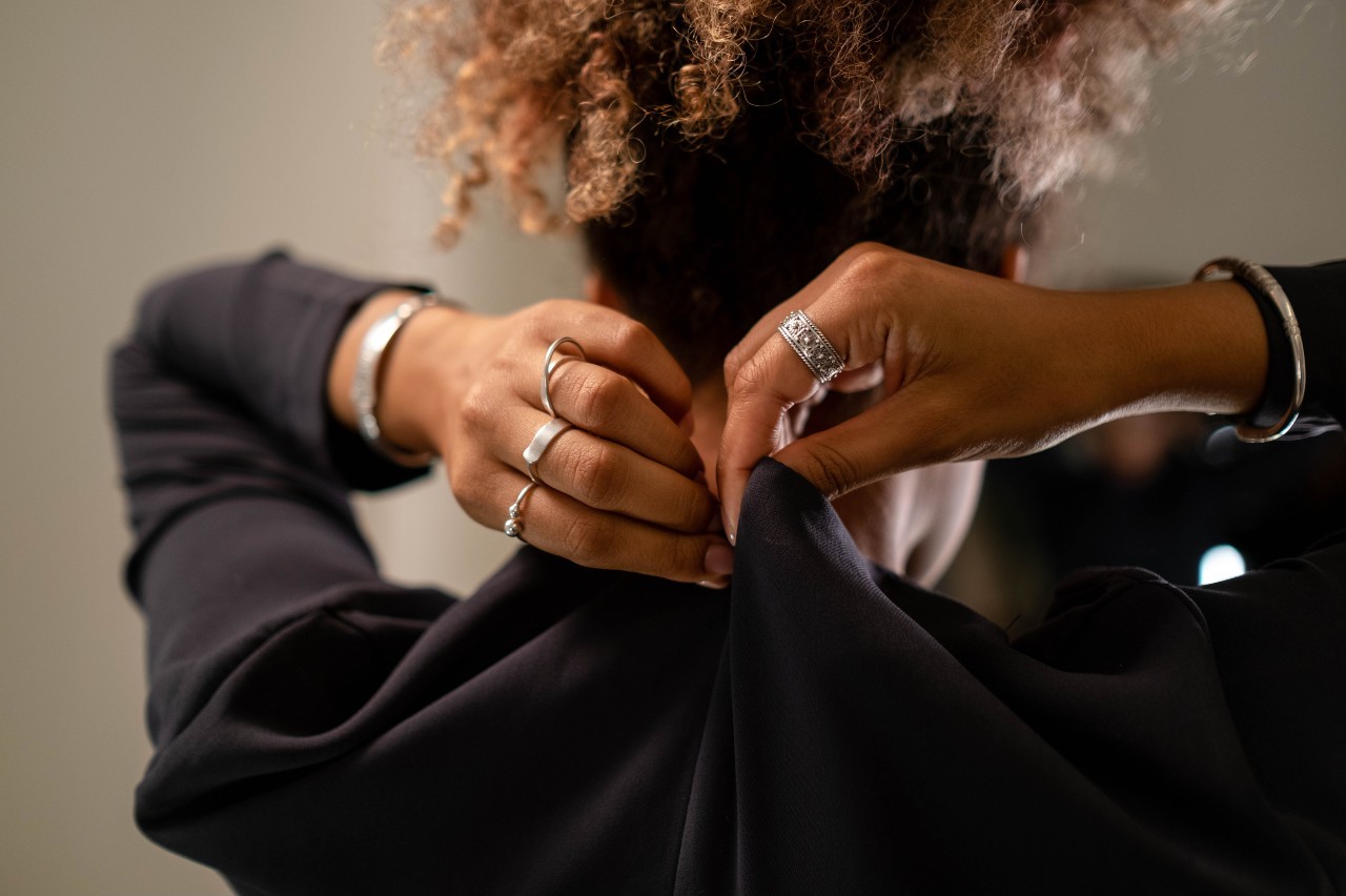 A close-up of a woman wearing a black jacket and white gold bangles and rings pinching and holding fabric from the back of her garment.