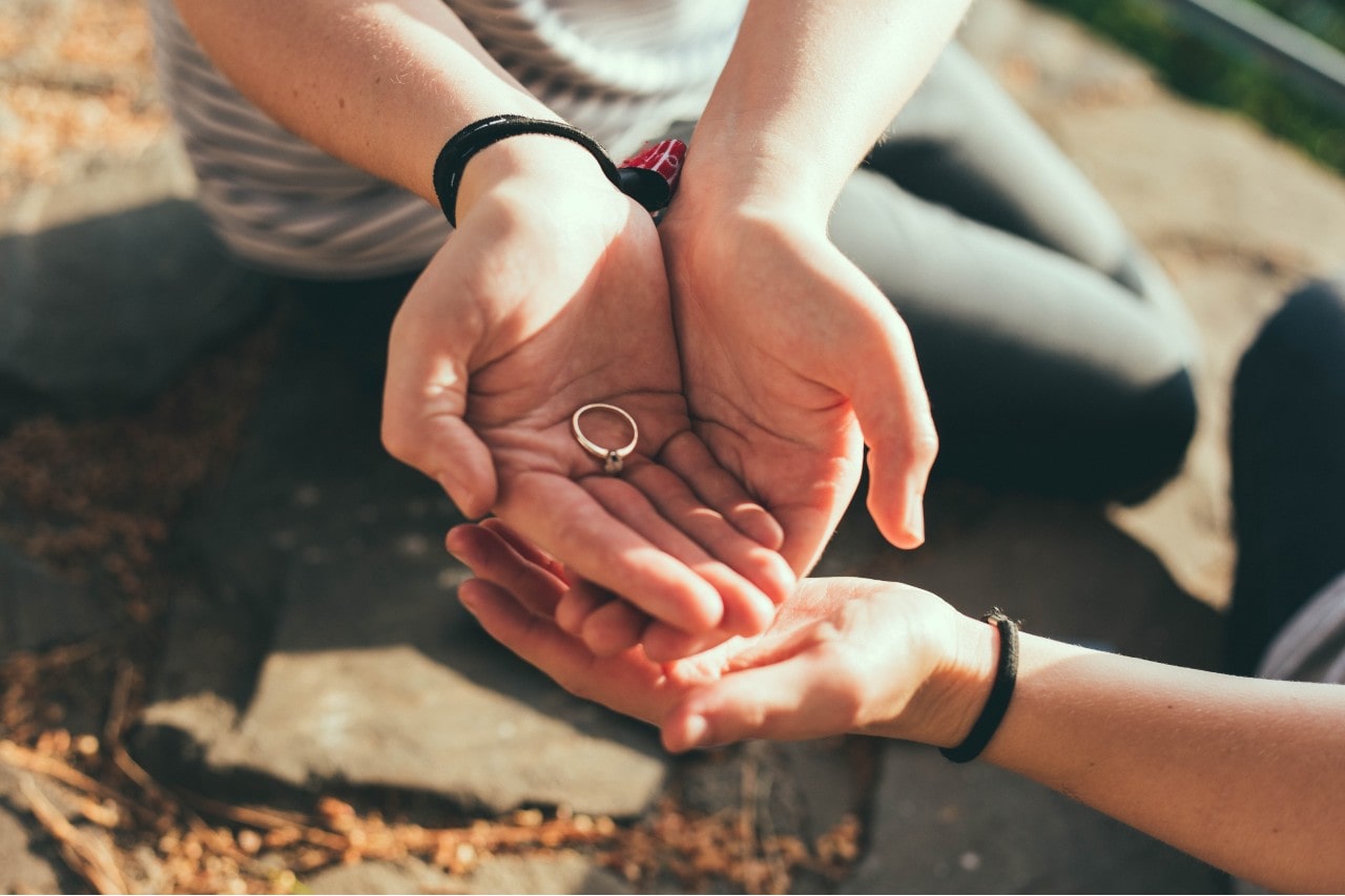 A man holds a solitaire gold marquise engagement ring while a woman’s hand supports him.