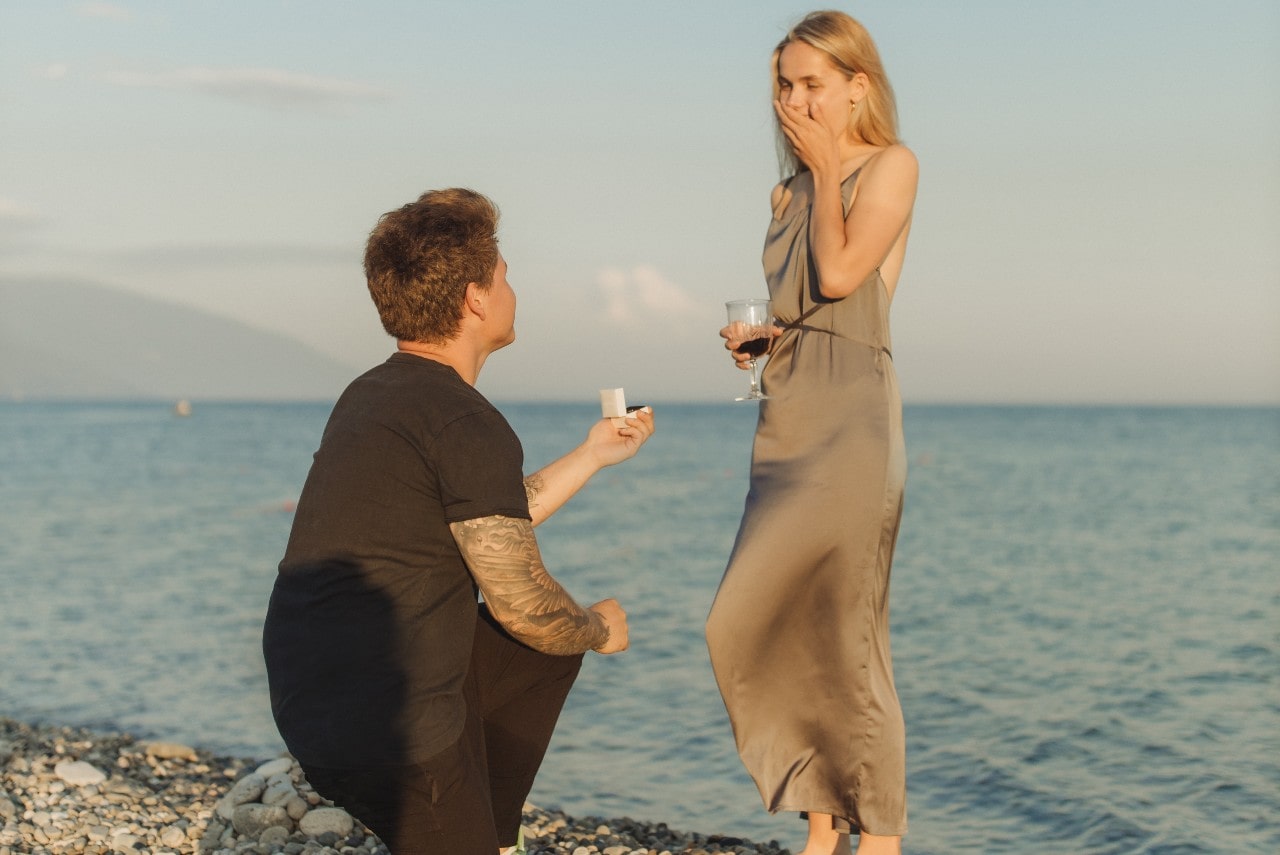 A tattooed man proposes to his girlfriend during sunset on a beach.
