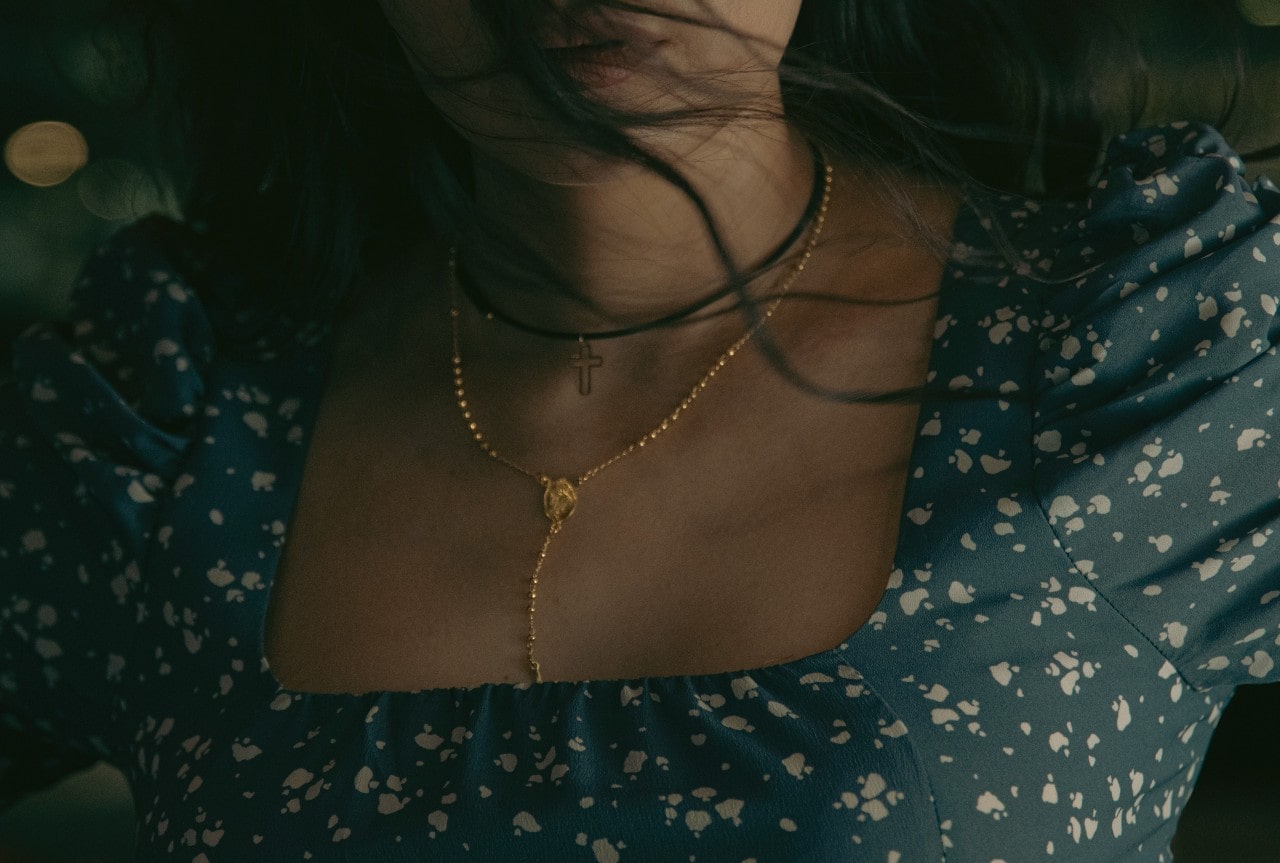 A woman wearing a blue patterned dress is shown with her hair blowing softly as she displays layered gold and black necklaces, including a delicate cross pendant.
