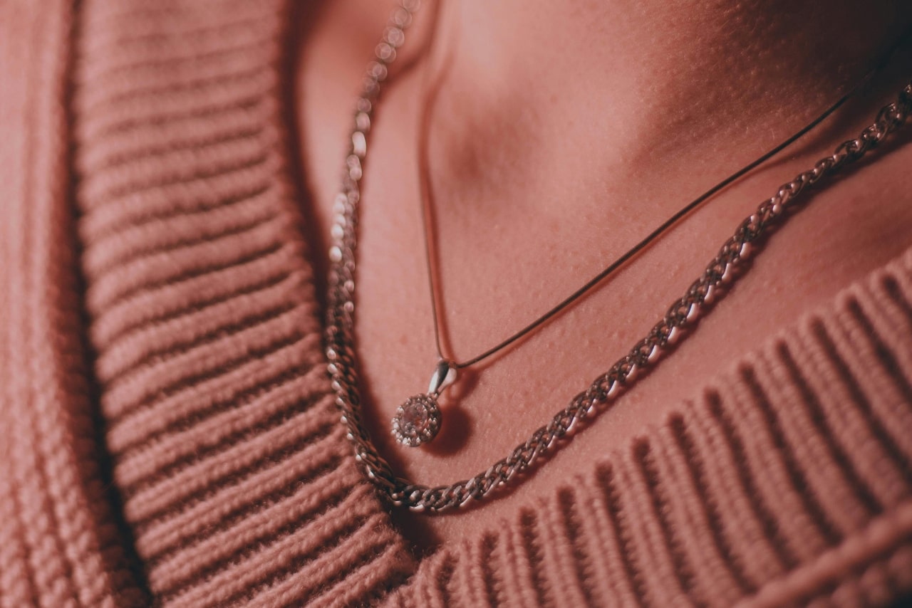 A close-up of a person wearing a halo diamond pendant necklace layered with a thicker chain over a ribbed v-neck sweater.
