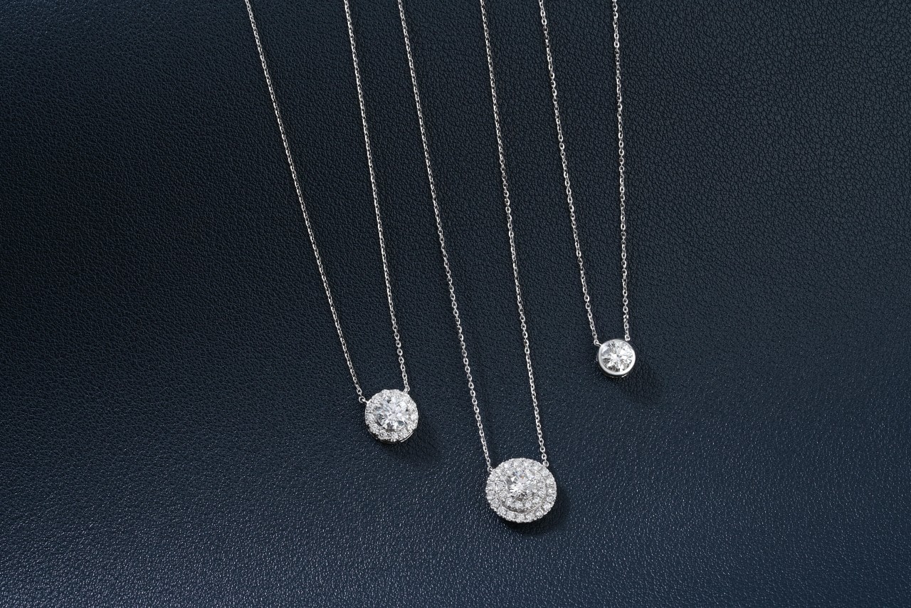 Three white gold necklaces with halo diamond pendants displayed on a dark textured background.