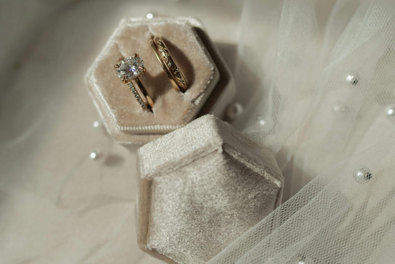 A wedding bridal ring set inside a beige velvet box on top of a white surface and near a veil with pearls. A wedding bridal ring set inside a beige velvet box on top of a white surface and near a veil with pearls.