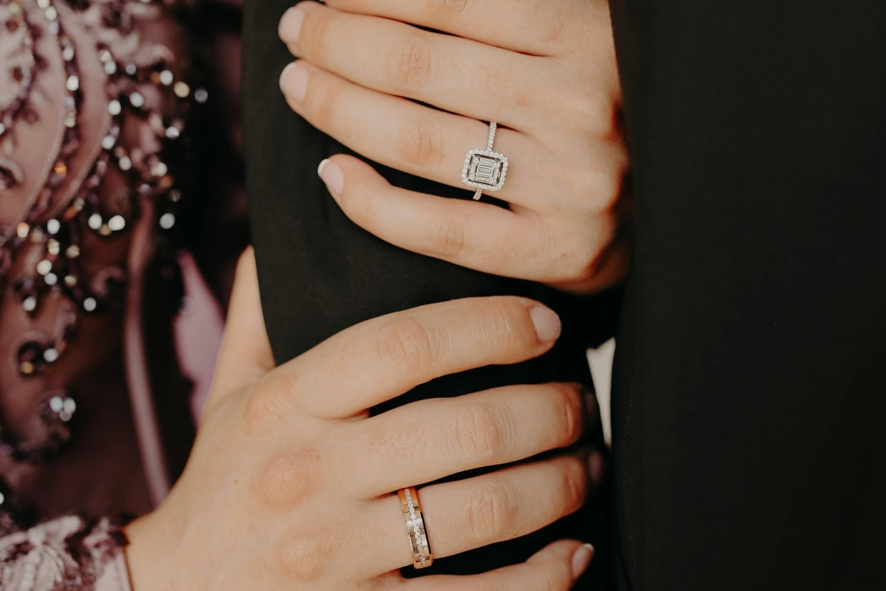 A close-up of a woman's hands held around a man’s arm, showcasing her diamond halo engagement ring. A close-up of a woman's hands held around a man’s arm, showcasing her diamond halo engagement ring.