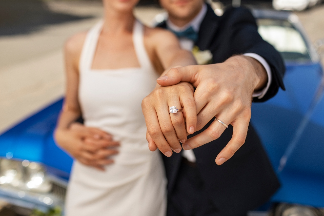 A newlywed couple in front of a blue car showcasing a diamond engagement ring on the bride’s hand. A newlywed couple in front of a blue car showcasing a diamond engagement ring on the bride’s hand.