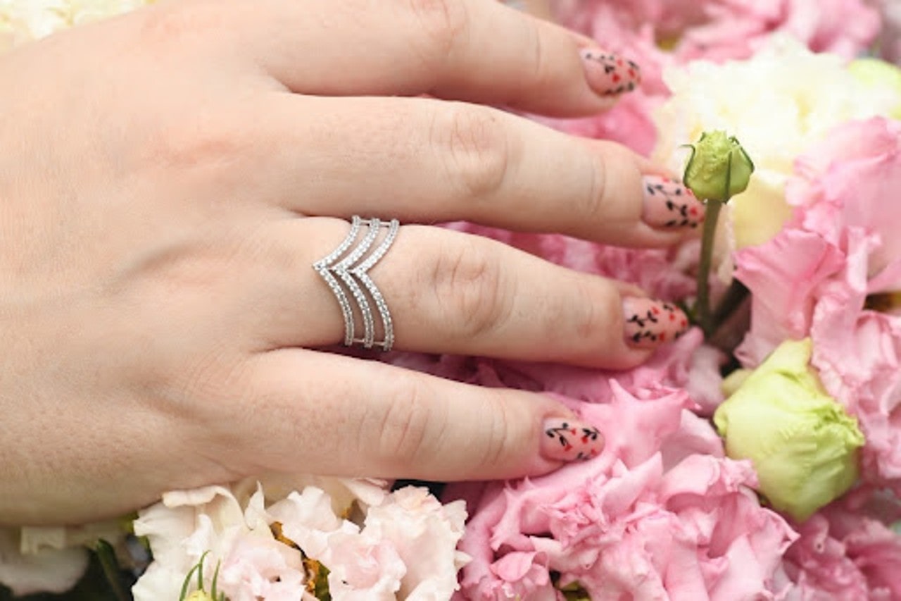 A hand with floral nail art wears a multi-band silver ring adorned with small diamonds, resting on a bed of pink and white flowers. A hand with floral nail art wears a multi-band silver ring adorned with small diamonds, resting on a bed of pink and white flowers.