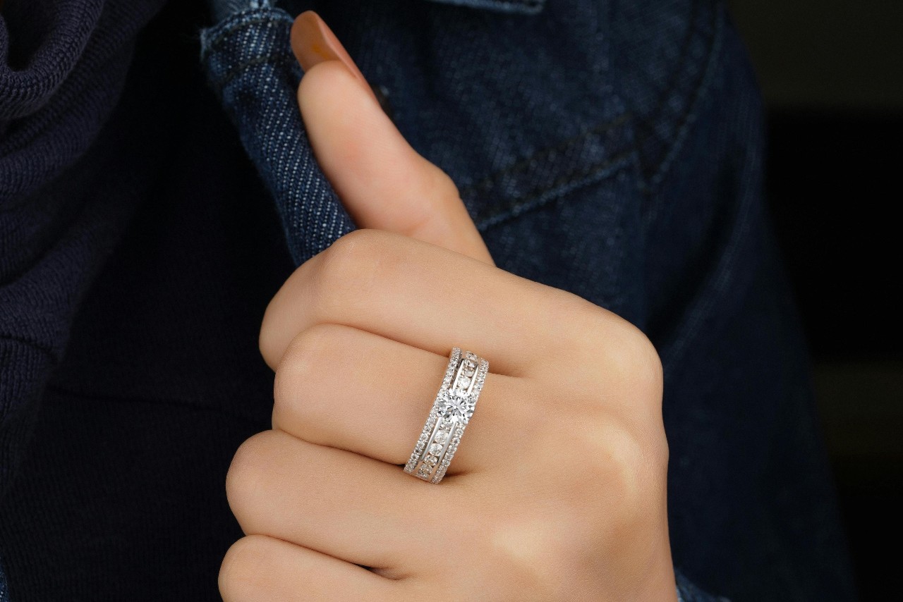 A person wearing a sparkling diamond wedding band on their finger with a denim jacket visible in the background. A person wearing a sparkling diamond wedding band on their finger with a denim jacket visible in the background.