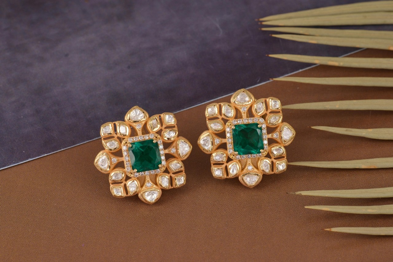 A close-up of a distinctive pair of yellow gold and diamond earrings with cushion cut emeralds at their centers. A close-up of a distinctive pair of yellow gold and diamond earrings with cushion cut emeralds at their centers.
