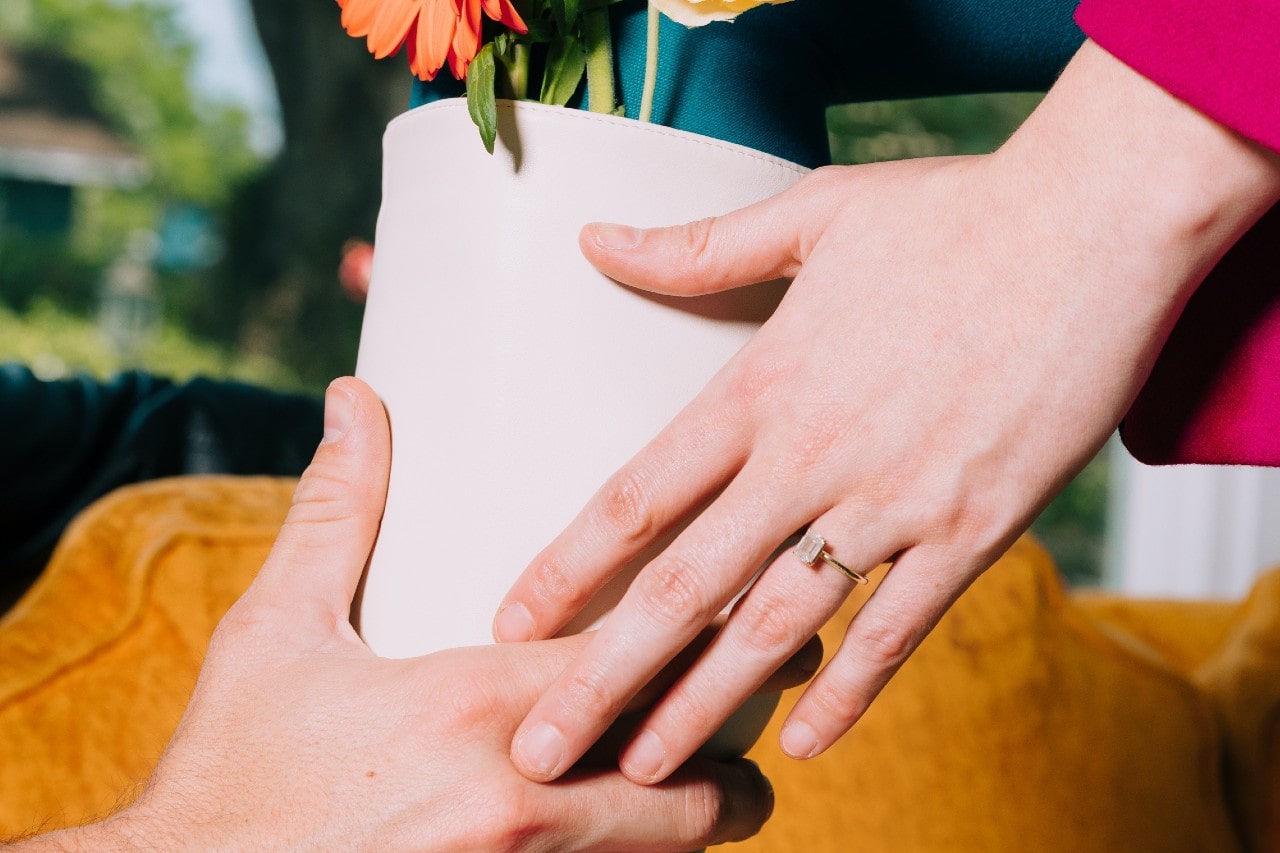 A man passes a potted plant to a woman wearing an emerald-cut solitaire ring.