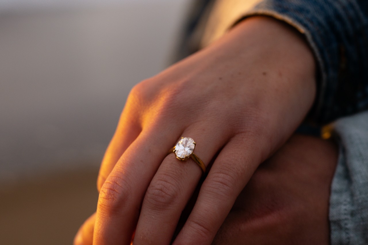 A woman’s hand wearing an oval-cut engagement ring and resting on a man’s hand.