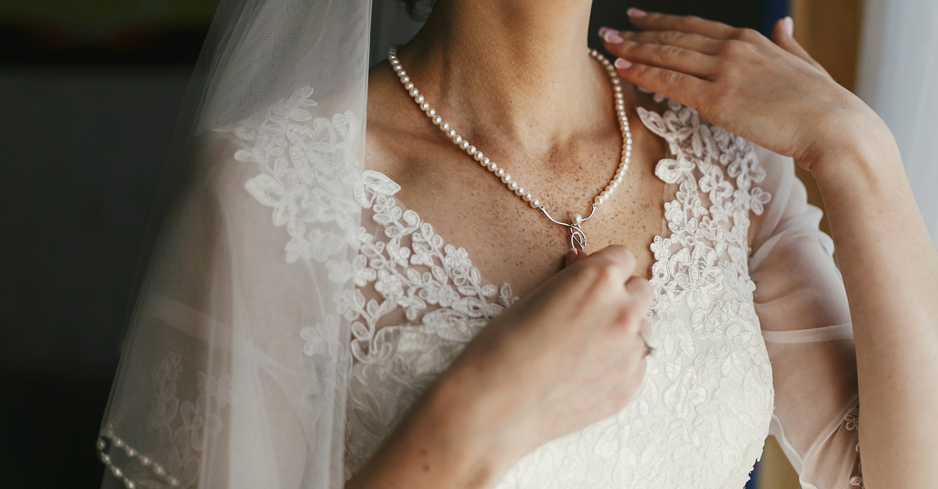 A bride touches her lace-trimmed wedding dress while wearing a classic pearl necklace. A bride touches her lace-trimmed wedding dress while wearing a classic pearl necklace.