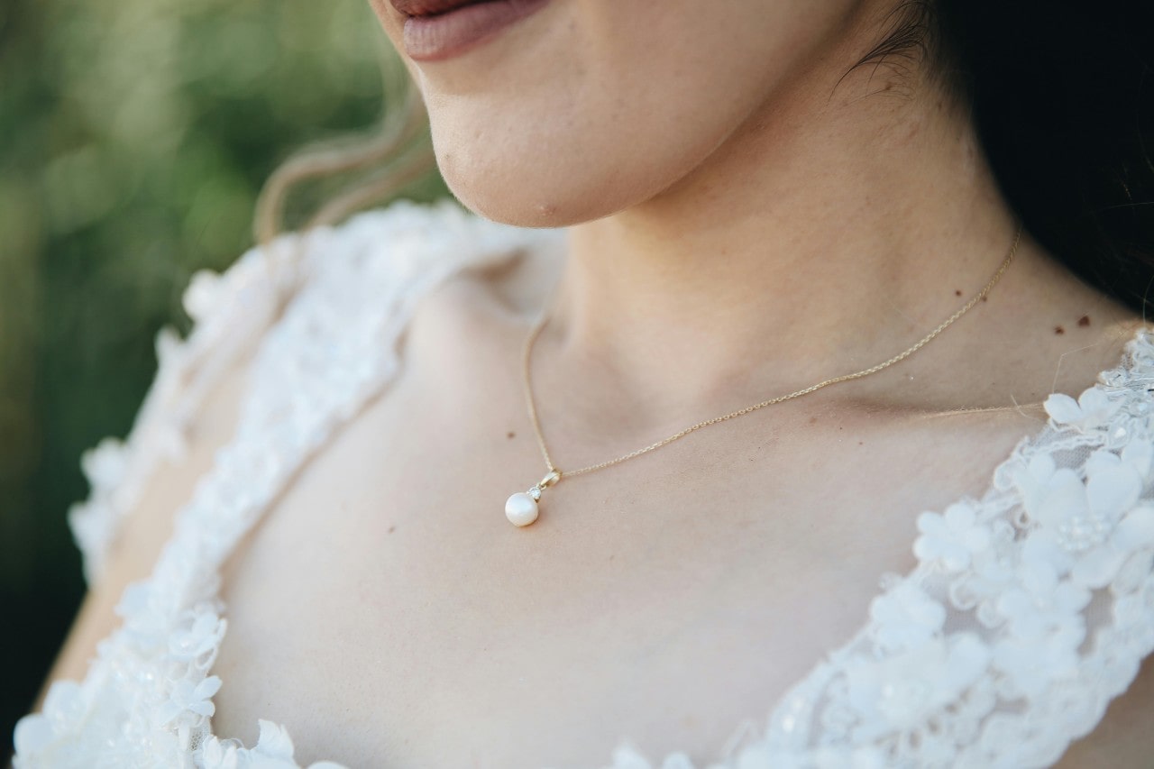 A close-up of a woman wearing a gold necklace with a single pearl pendant, dressed in a white lace dress. A close-up of a woman wearing a gold necklace with a single pearl pendant, dressed in a white lace dress.